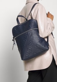 Sac à dos bleu marine en matériau synthétique texturé avec logo en relief, une fermeture zippée en haut et des bretelles réglables.