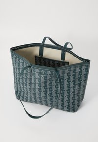 Zelená taška na rameno so vzorom v geometrickom dizajne, s uzatváraním na zips a dvoma dlhými rúčkami; vnútro je v kontrastnej svetlej farbe.