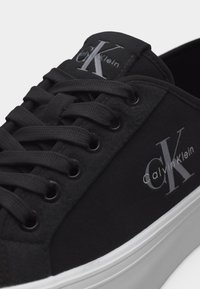 Baskets Calvin Klein noires avec des lacets noirs, logo blanc "CK" sur le côté et la languette, et semelle en caoutchouc blanche.