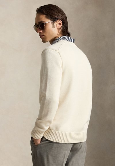 Jeune homme portant des lunettes de soleil, un pull en tricot crème sur une chemise rayée à col avec un pantalon gris, debout les mains dans les poches.