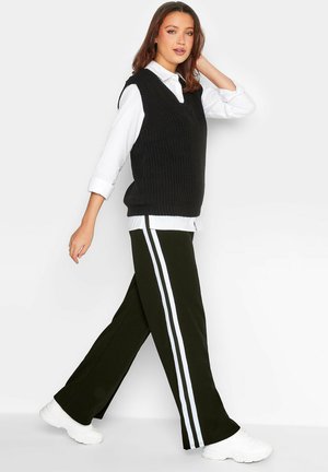 Svart stickad ärmlös tröja över en vit skjorta med krage, kombinerad med svarta byxor med vita sidostripes och vita sneakers.