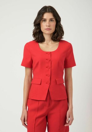 Vrouw met donker schouderlang haar, gekleed in een rode kortgeknoopte jas en bijpassende rode broek, staand tegen een effen witte achtergrond.