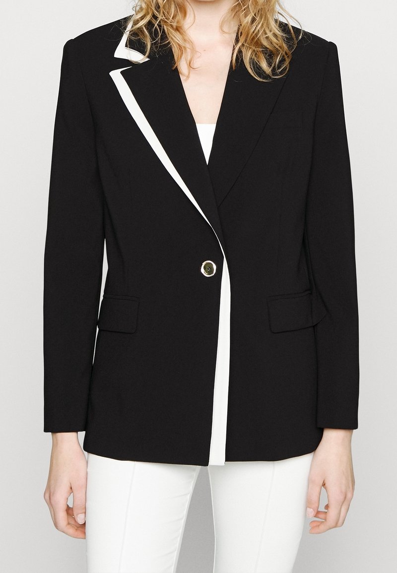 Femme portant un blazer noir avec un bord blanc sur le revers, boutonné au centre, assorti à un pantalon blanc et un haut intérieur blanc.