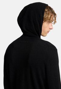 Felpa nera in maglia con una texture morbida, caratterizzata da un design semplice, dotata di cappuccio e di un'etichetta del marchio discreta sulla parte posteriore.