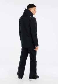 Zwarte waterdichte jas met een capuchon, ritssluitingen aan de zijkanten en een hoge kraag, gecombineerd met een zwarte broek en stevig schoeisel.