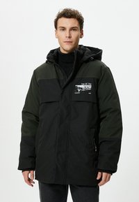 Schwarze und dunkelgrüne gepolsterte Jacke mit Kapuze, ausgestattet mit einem Frontreißverschluss, einem Logo-Patch auf der Brust und seitlichen Taschen. Strukturierter Stoff mit glatter Oberfläche.