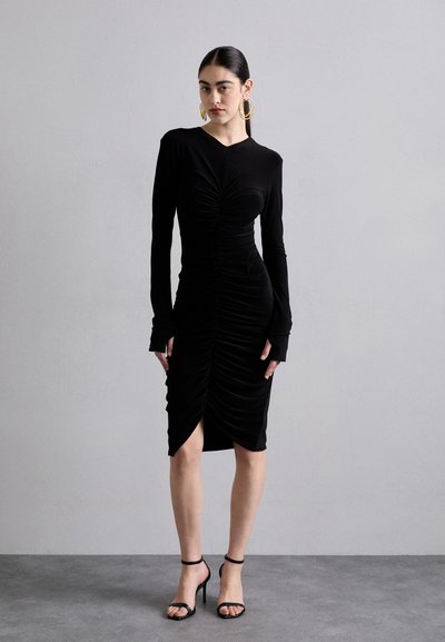 Black, long-sleeve dress with ruched texture, a deep neckline, and side slit. Paired with black strappy heels and large hoop earrings.
