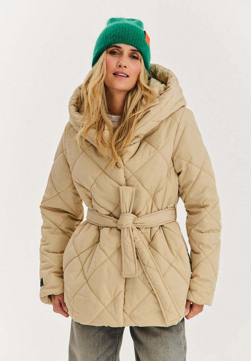 Žena v béžovém prošívaném kabátu s opaskem a zelené pletené čepici, stojící před jednoduchým bílým pozadím.