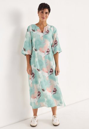 Frau steht und trägt ein locker sitzendes Midikleid mit pastellfarbenem Aquarellmuster, 3/4-Ärmeln und weißen Turnschuhen vor weißem Hintergrund.