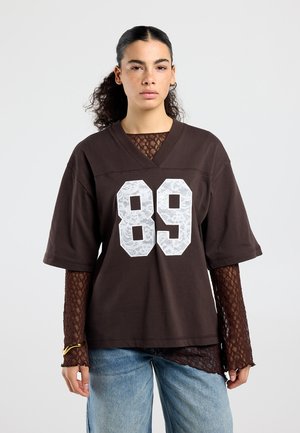 Jonge vrouw met krullend haar draagt een bruine oversized blouse met een witte "89", een kanten top met lange mouwen eronder, een blauwe spijkerbroek en een gouden armband aan haar linkerpols.
