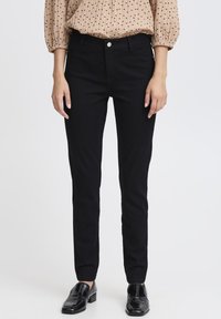 Pantalons slim noirs en denim, avec un design classique à cinq poches et une fermeture à bouton. Portés avec un blouse beige texturé.