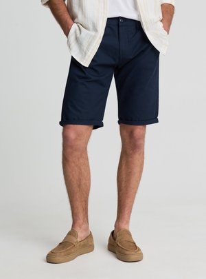 Uomo con pantaloncini blu navy risvoltati, scarpe beige slip-on, camicia bianca e giacca aperta beige chiaro, in piedi con le mani nelle tasche.