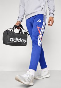 Zili sporta bikses ar baltām un sarkanām akcentiem, ar trim baltām svītrām sānos. Melna sporta soma ar baltu "adidas" logotipu tiek turēta rokā.