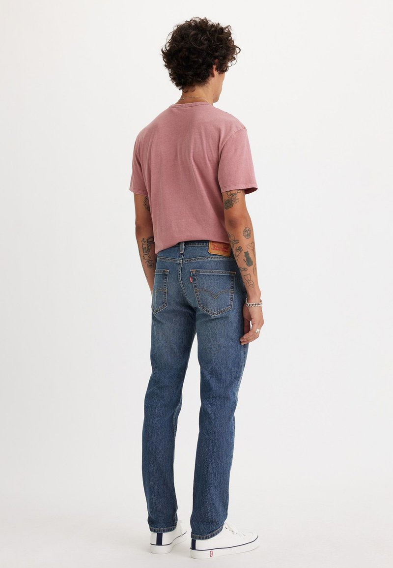 T-shirt rosa chiaro a maniche corte abbinato a jeans di denim blu. I jeans presentano un taglio classico, tasche posteriori e un dettaglio in pelle marrone. Sneakers bianche.
