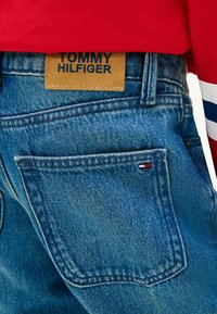 Jeans de mezclilla azul claro con un prominente parche de cuero etiquetado "TOMMY HILFIGER" en la cintura, que presenta un pequeño detalle bordado de la bandera en el bolsillo trasero.