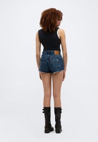 Γυναίκα με σγουρά μαλλιά στέκεται με την πλάτη γυρισμένη, φορώντας μαύρο αμάνικο τοπ, ξεφτισμένο τζιν σορτς και μαύρες μπότες μέχρι τη μέση της γάμπας με αγκράφες.