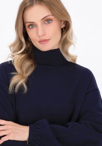 Granatowy sweter z golfem o prążkowanej teksturze, luźnych rękawach i subtelnym kształcie balonu; modelka ma luźne, falowane włosy i złote kolczyki w kształcie kół.