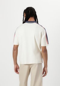 Polo din tricot, cu mânecă scurtă, crem, cu guler bleumarin și accente roșii pe umeri. Are o croială lejeră și margine ribb.