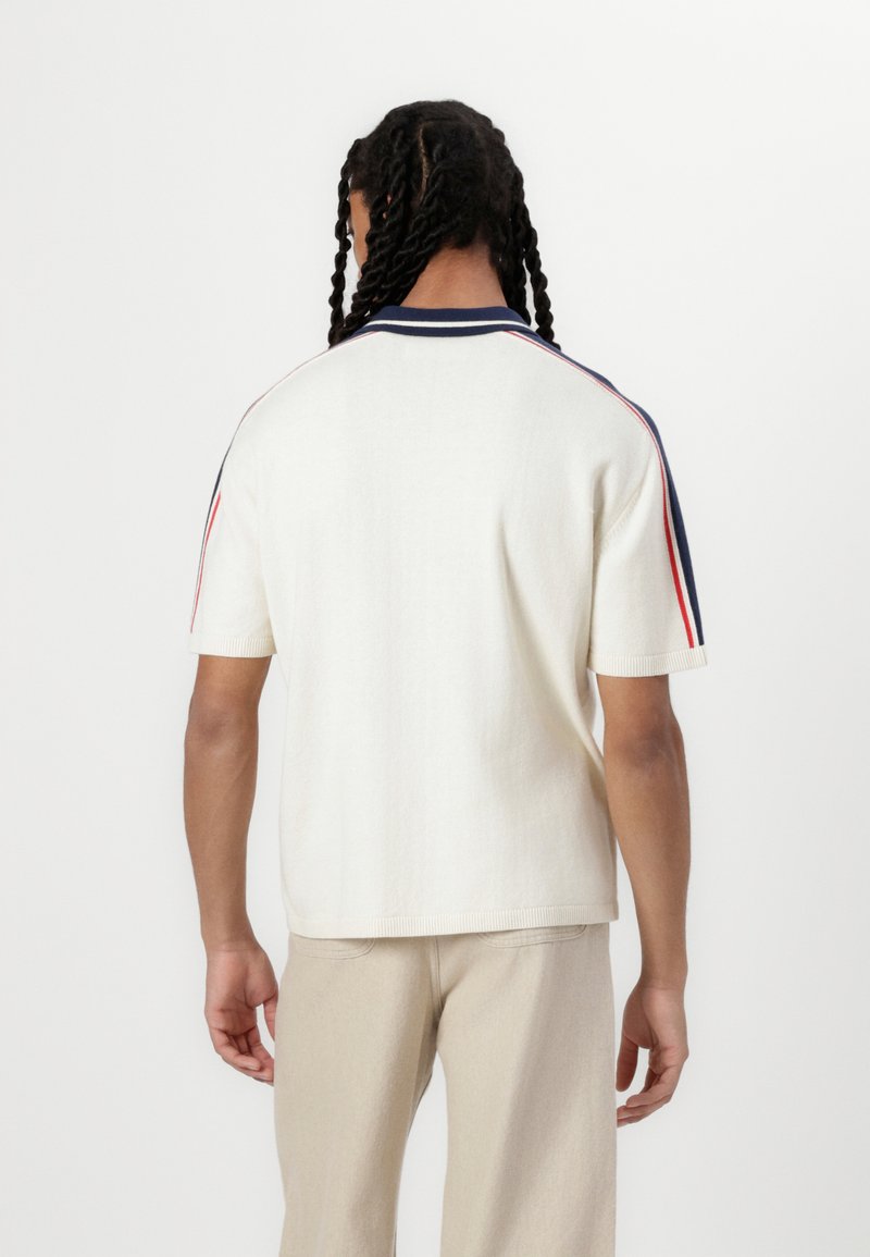 Polo de punto de manga corta color crema con cuello azul marino y acentos rojos en los hombros. Presenta un corte relajado y dobladillo acanalado.