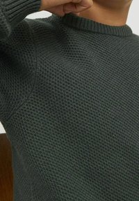 Närbild av en person som bär en mörkgrön, texturerad stickad tröja med ribbad krage, som höjer en arm nära sitt ansikte.