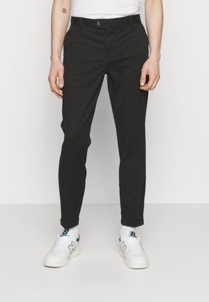 Mann trägt schwarze schmale Hose, weiße Sneaker mit grünen Akzenten und ein weißes Hemd, steht vor schlichtem Hintergrund.
