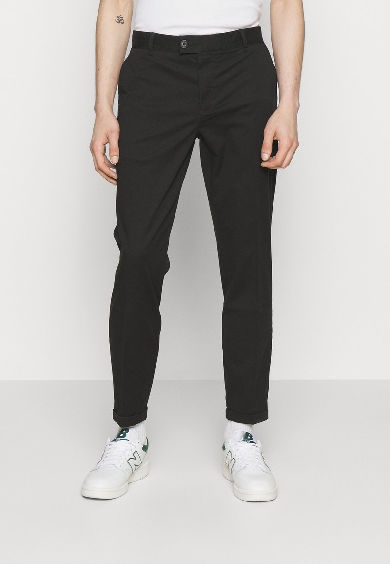 Homme portant un pantalon noir slim, des baskets blanches avec des accents verts, et une chemise blanche, debout devant un fond uni.
