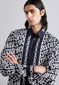 Μαυρόασπρο πουκάμισο με σχέδιο, που διαθέτει έντονο κείμενο και σχέδια με αλυσίδες, κουμπάκρι μπροστά, μακριά μανίκια και λεία υφή υφάσματος.