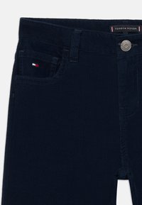 Navy corduroy pants with a ribbed texture, featuring a silver button, front pocket, and a small flag logo on the pocket.