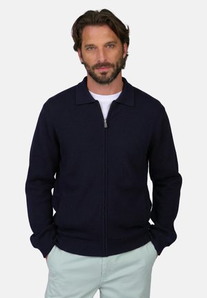 Homme barbu portant un pull à fermeture éclair bleu marine et un pantalon de couleur claire, debout les mains dans les poches devant un fond uni.