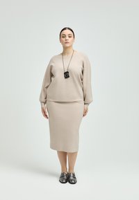 Beige gestrickter Pullover mit übergroßen Ärmeln, kombiniert mit einem taillierten beigen Rock. Die Accessoires umfassen eine schwarze Halskette mit Anhänger und dunkle Loafers.