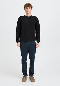 Melnas adītas džemperis ar apaļu kakla izgriezumu, uzvilkts virs tumši zilām džinsu biksēm. Aksesuāri ietver brūnus kurpes ar saišu stiprinājumu. Vienkāršs un ikdienišķs dizains.