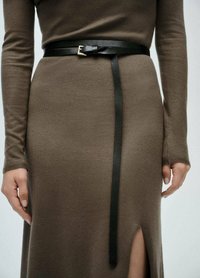 Cintura in pelle nera con fibbia argentata, indossata sopra un abito lungo a maniche lunghe in maglia marrone, con uno spacco alto fino alla coscia sul lato.