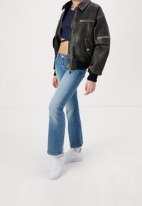 Chaqueta bomber de cuero negro con bolsillos con cremallera, llevada sobre un top corto oscuro. Combinada con jeans de campana de color azul claro y zapatillas blancas.