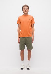 Νεαρός άνδρας που στέκεται, φοράει πορτοκαλί κοντομάνικο πουκάμισο, χακί σορτς, λευκές κάλτσες και λευκά αθλητικά παπούτσια μπροστά σε απλό λευκό φόντο.