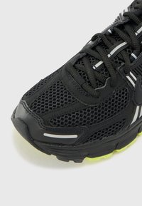 Chaussure de sport noire avec une tige en maille, semelle en caoutchouc, rayures blanches contrastantes et un accent jaune vif sur le talon.
