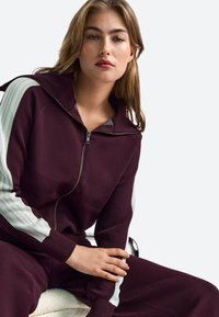 Burgundska majica s kapuco na zadrgo s spadajočimi hlačami; ima teksturirano tkanino, kontrastne svetlo zelene črte na rokavih in prostorno kapuco.