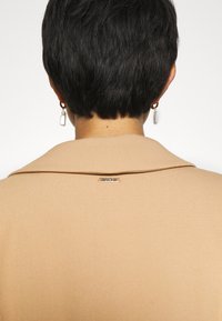 Cappotto beige con colletto a scollo, texture morbida e una piccola etichetta metallica sul retro, indossato con orecchini a perla pendenti.