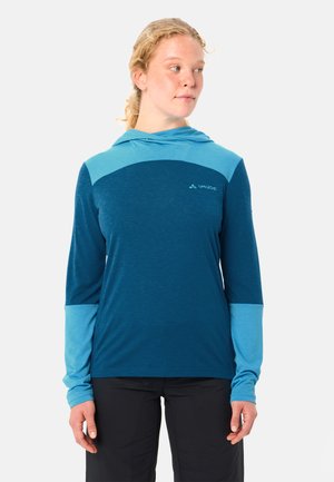 Donna con capelli chiari che indossa una felpa con cappuccio bicolore blu a manica lunga e pantaloni neri, che guarda leggermente verso destra.