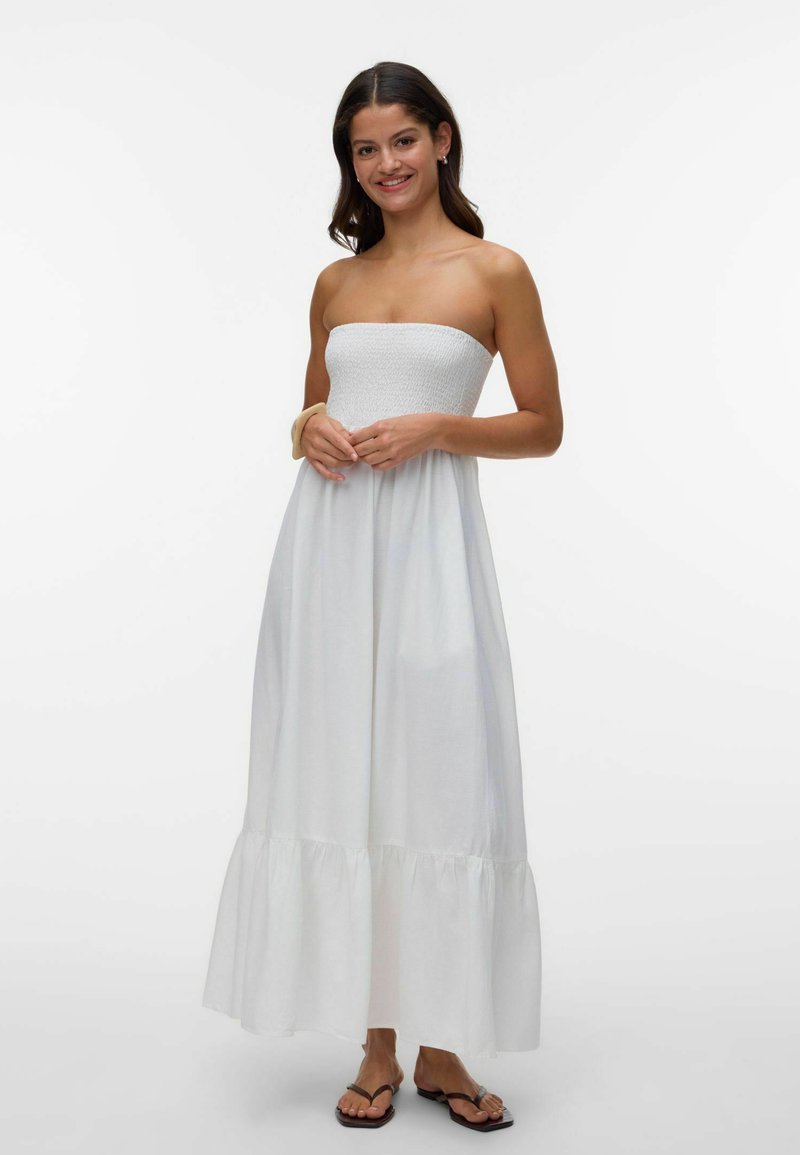 Tânără zâmbitoare, purtând o rochie maxi albă fără bretele și papuci negri, stând cu mâinile ușor încleștate.