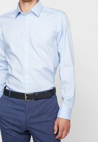 Lichtblauw button-up shirt met een puntige kraag, met een subtiele textuur. Gecombineerd met donkerblauwe broek en een zwarte riem.