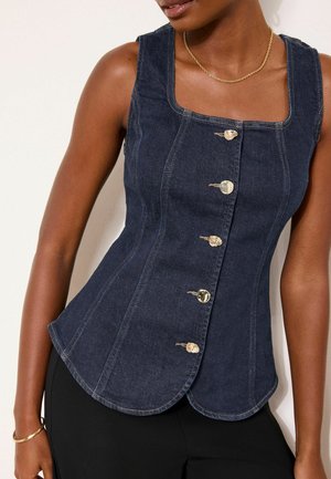 Frau trägt ein eng anliegendes, dunkelblaues, ärmelloses Denim-Top mit goldenen Knöpfen und geschwungenem Saum, kombiniert mit schwarzer Hose und goldschmuck.