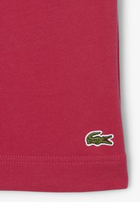 Camiseta de algodón roja con una textura suave y un logo de cocodrilo bordado en verde en la parte inferior izquierda. El dobladillo está perfectamente cosido.
