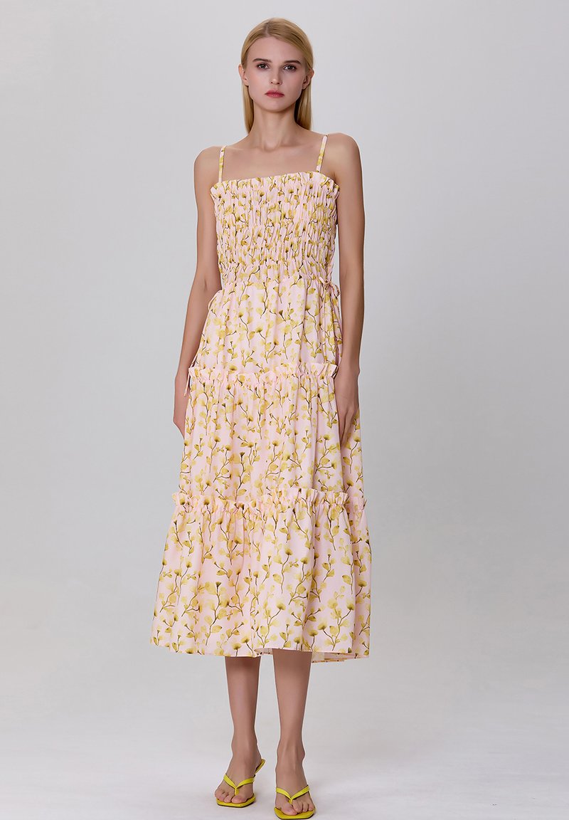 Robe midi à imprimé floral rose, avec un corsage froncé, des fines bretelles et des détails en volants superposés au niveau de l'ourlet. Portée avec des sandales jaunes.