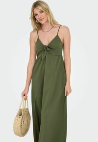 Robe maxi vert olive avec un détail noué à l'avant, de fines bretelles spaghetti et une silhouette fluide. Accompagnée d'un sac en paille tressée.