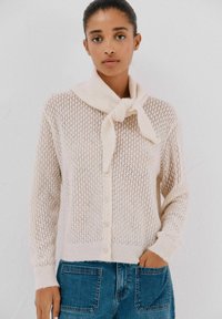 Cardigan di maglia beige chiaro con una trama larga e aperta, caratterizzato da un nodo sul colletto e chiusura con bottoni. Abbinato a jeans in denim blu.