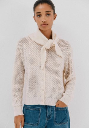 Licht beige gebreide cardigan met een losse, open weving, voorzien van een strik bij de hals en een knoopsluiting. Gecombineerd met blauwe spijkerbroek.