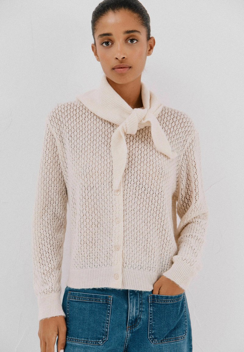 Cardigan di maglia beige chiaro con una trama larga e aperta, caratterizzato da un nodo sul colletto e chiusura con bottoni. Abbinato a jeans in denim blu.