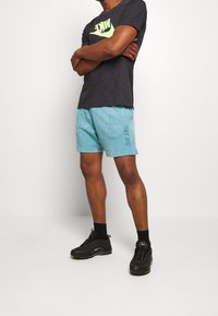 Man som står med armarna i kors, iklädd svart Nike-t-shirt, blå shorts med "Just Do It"-text, svarta strumpor och svarta Nike-sneakers.