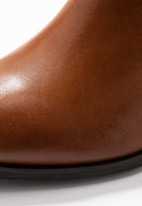 Ruskeat nahkaiset nilkkurit, joissa on sileä pinta, kaareva päällinen ja musta kumipohja. Päällinen on tasaista pintaa ilman näkyviä saumoja.