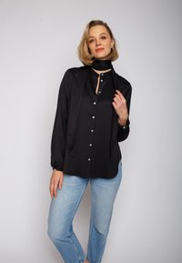 Czarna satynowa koszula na guziki z długimi rękawami, z detalem w postaci wiązania przy szyi i srebrnymi guzikami, zestawiona z jasnoniebieskimi dżinsami. Gładka faktura.